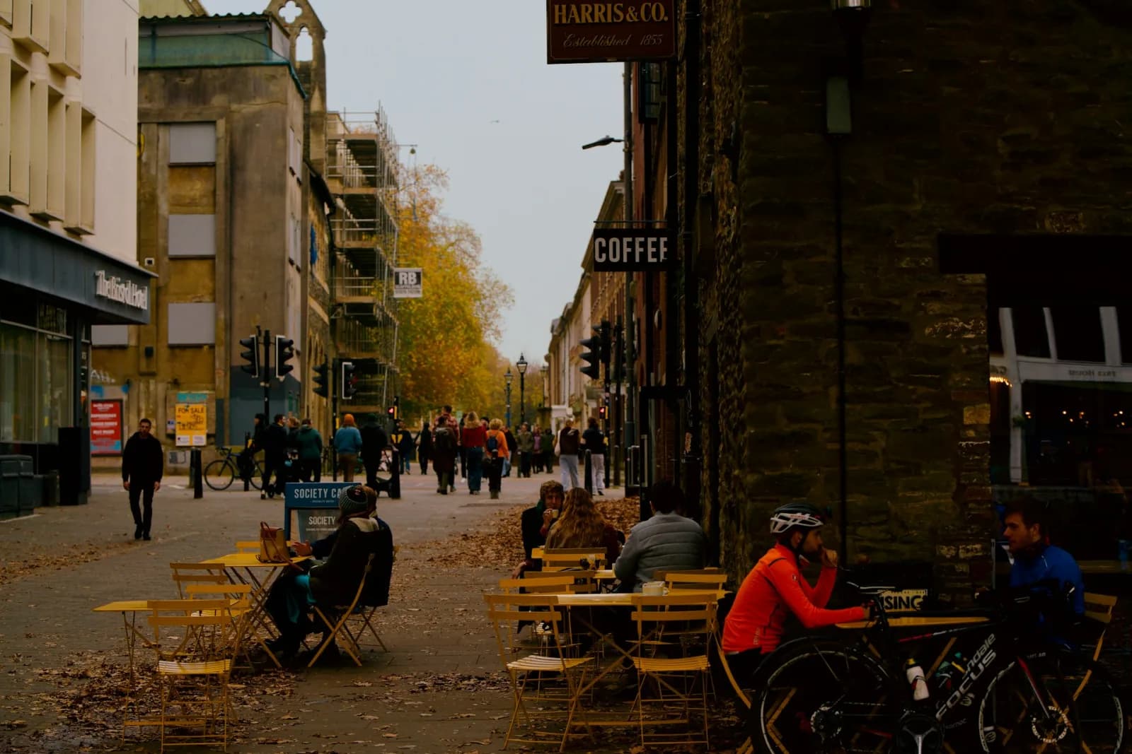 A person sitting at a small local cafe on a residential street, holding a coffee cup with other locals visible in the background