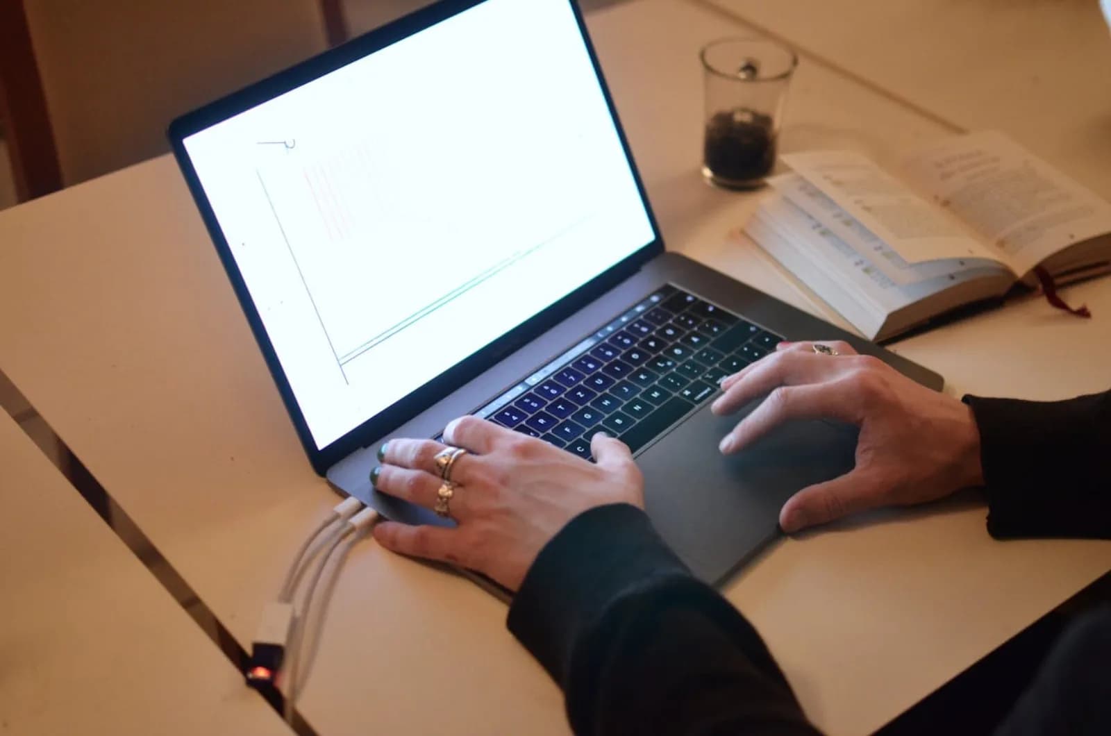 Close-up of hands typing on a laptop at a plain desk, with an ethernet cable and a second screen showing a code editor in the background