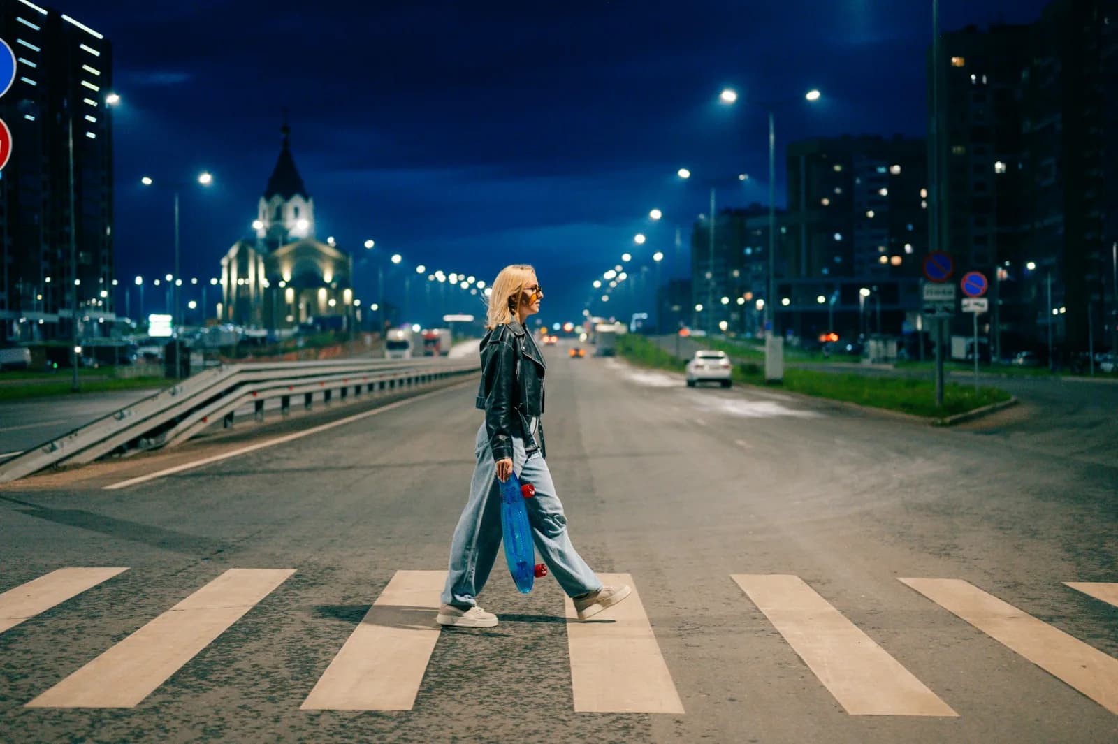 A woman walking alone on a well-lit city street in the evening, passing local shops with other pedestrians in the background
