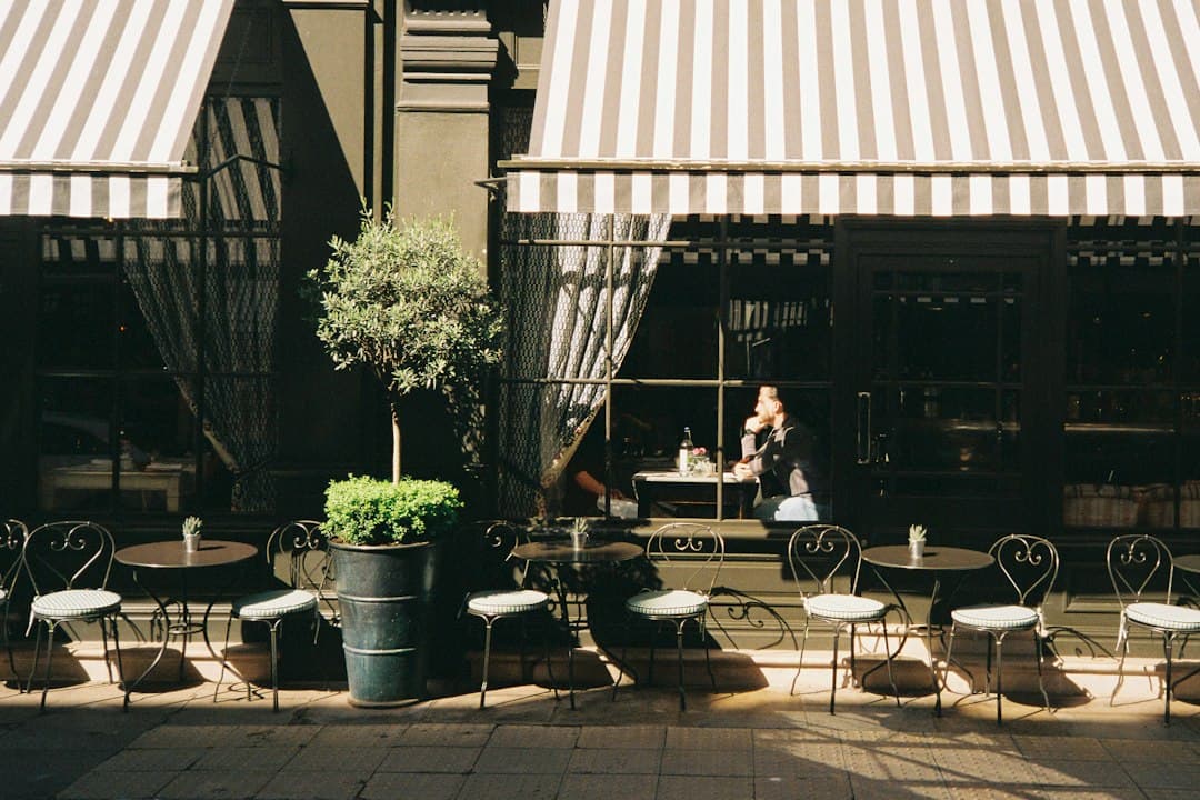 Outdoor café terrace with a striped awning, tables, and a person working on a laptop in the sunshine.