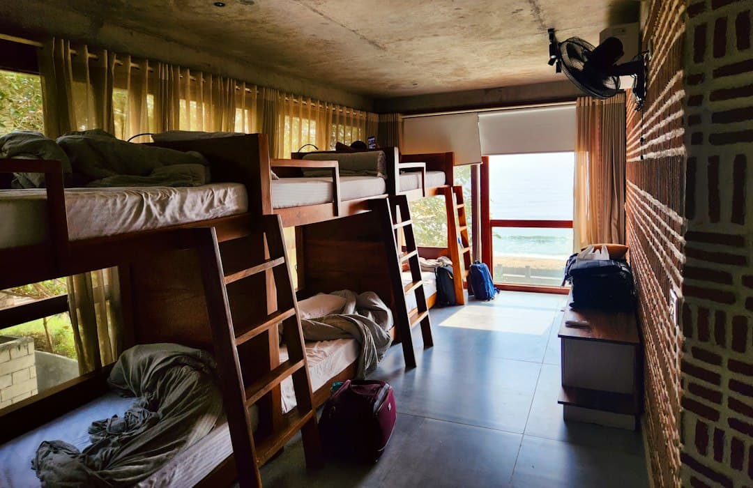 A sunlit hostel dormitory room with bunk beds and luggage, featuring a view of the ocean.