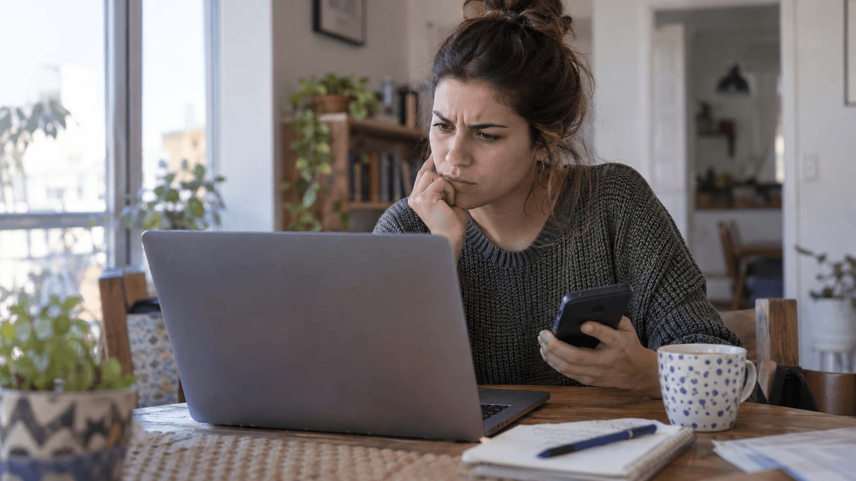 Woman at a home desk using a laptop and phone to access digital administrative information.