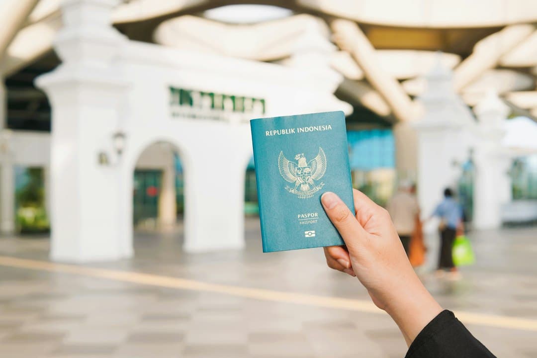 A hand holding an Indonesian passport in front of an airport terminal entrance.