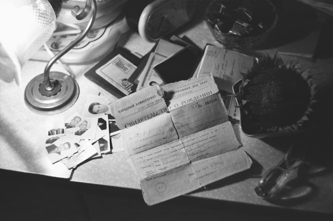 Administrative documents and a passport spread out on a desk under a lamp, top-down view in black and white.