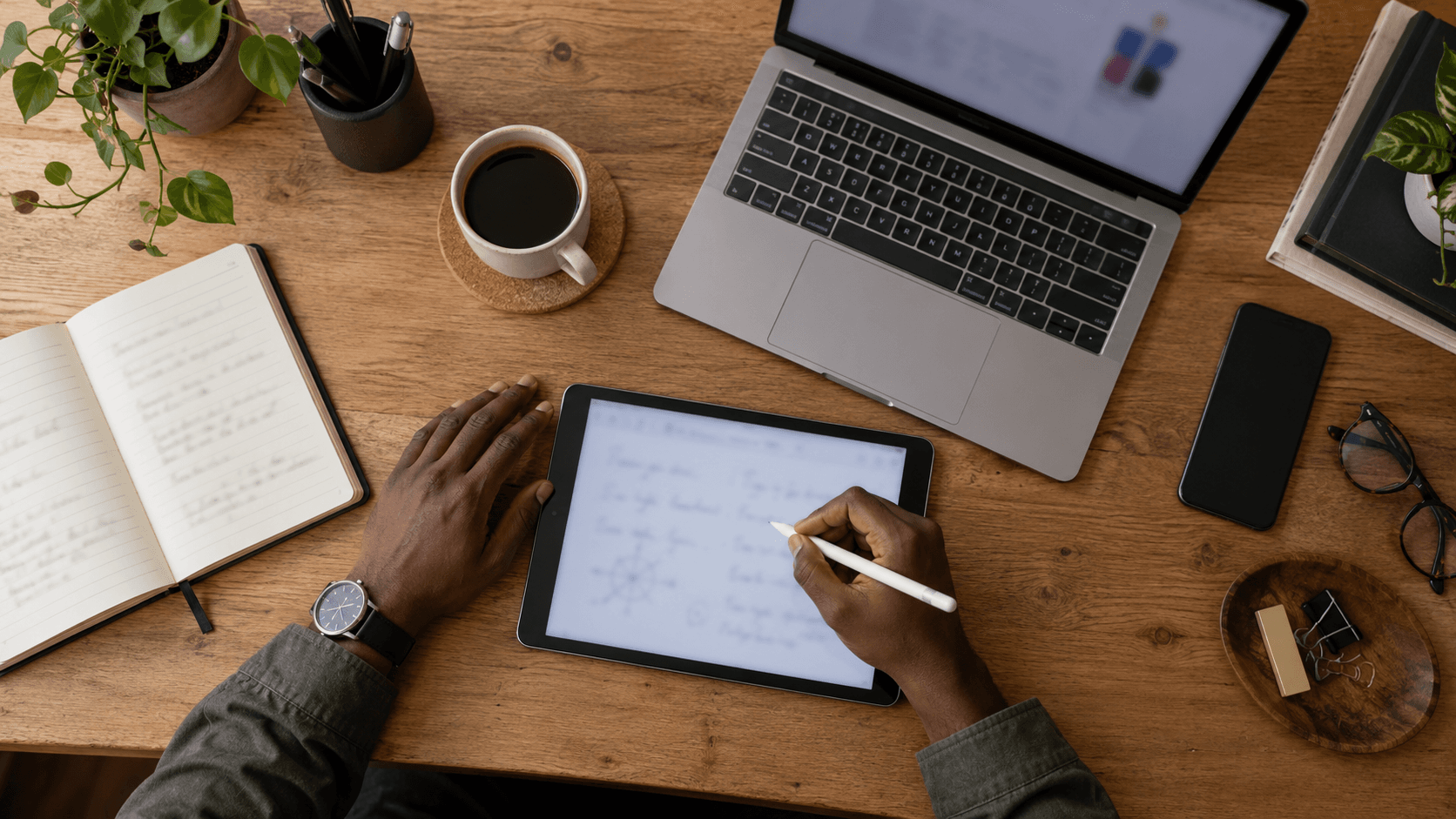 Digital nomad workspace with laptop, tablet, coffee and notebook on a wooden desk.