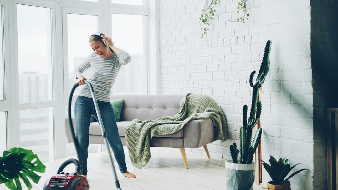 Woman vacuuming a bright living room with a sofa, plants, and large windows.