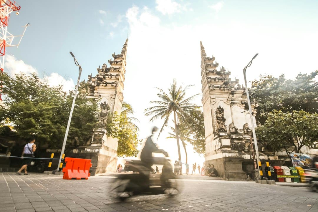 Conducteur de scooter en mouvement près d'un temple balinais avec palmiers et portes traditionnelles.