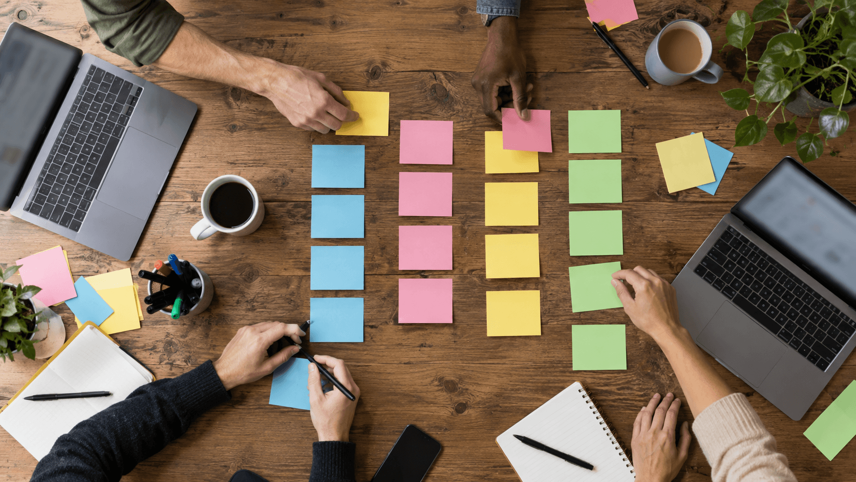 Espace de travail collaboratif avec ordinateurs portables, post-its et café sur table en bois avec plusieurs travailleurs.