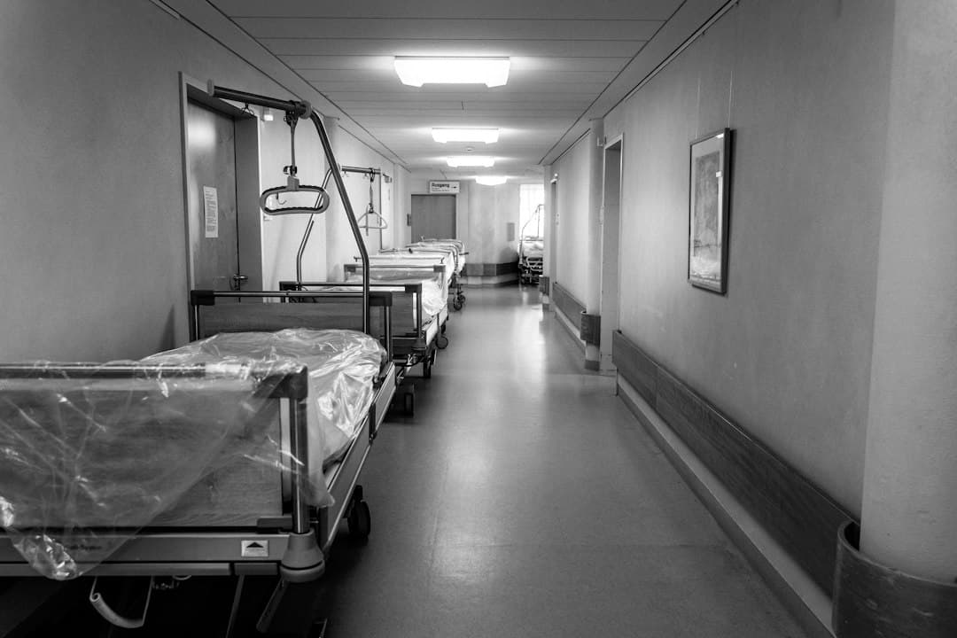 Empty hospital corridor with medical beds and fluorescent lighting, black-and-white photograph.