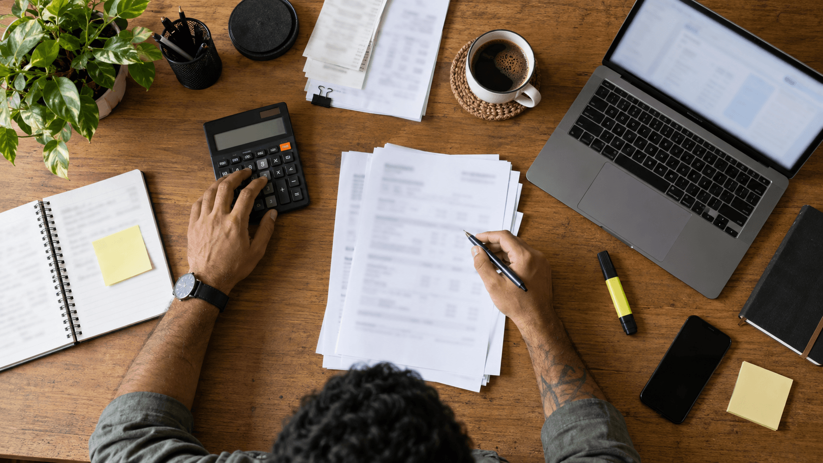 Person at a desk with a calculator, tax documents, laptop, and coffee reviewing papers.