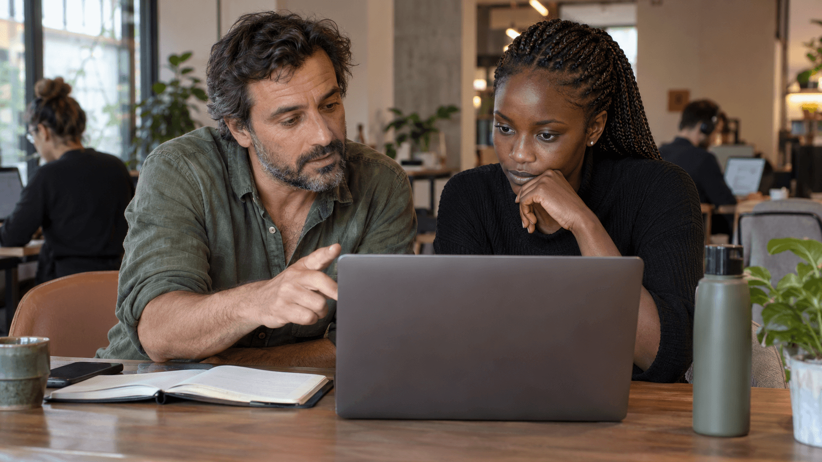 Two professionals collaborating on a laptop at a coworking table with plants and colleagues nearby.