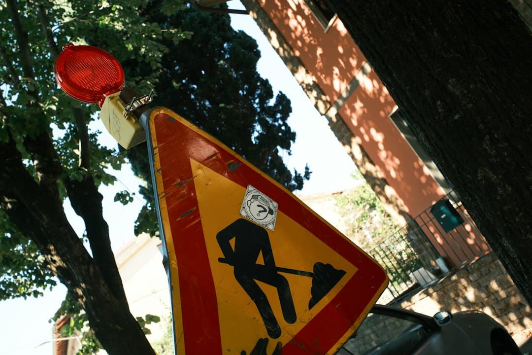 A construction warning sign with a blinking light and a sticker, in front of a tree and a building.