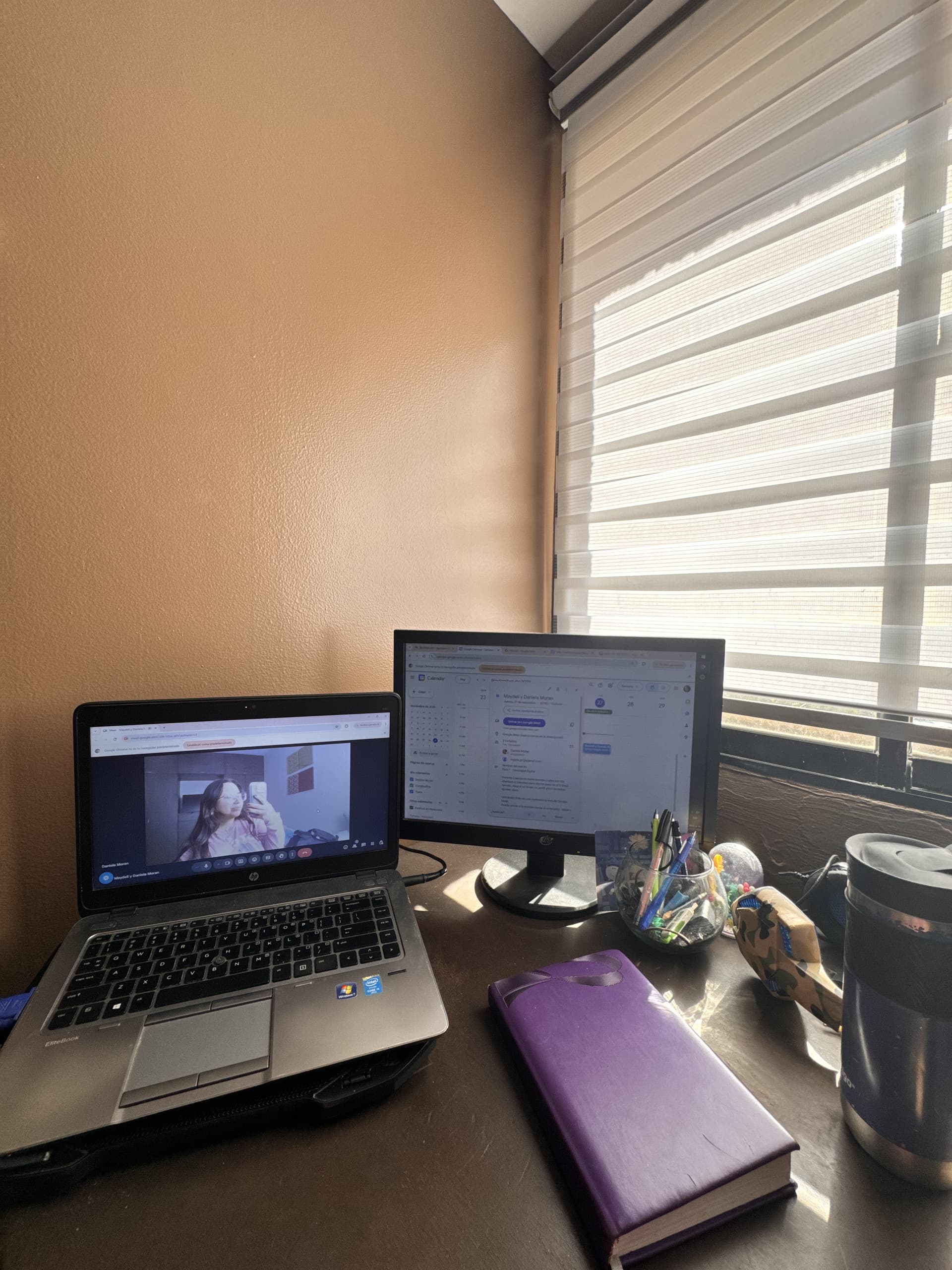 A home office with a laptop on a video call and an external monitor displaying a calendar, near a window.