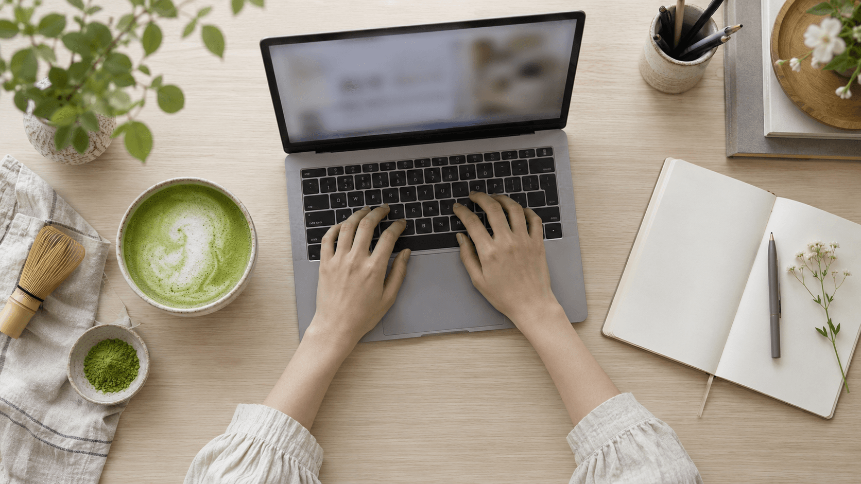 Overhead view of an organized workspace with a laptop, notebook, matcha drink and plants on a light wooden desk.