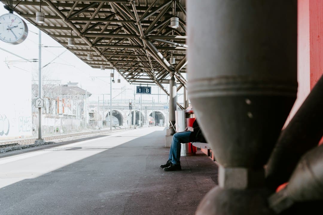 A traveler sitting on a bench on a train platform, working on their laptop while waiting.