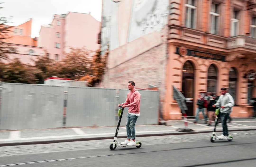 Person riding an electric scooter on an urban street with historic architecture and another visible rider.