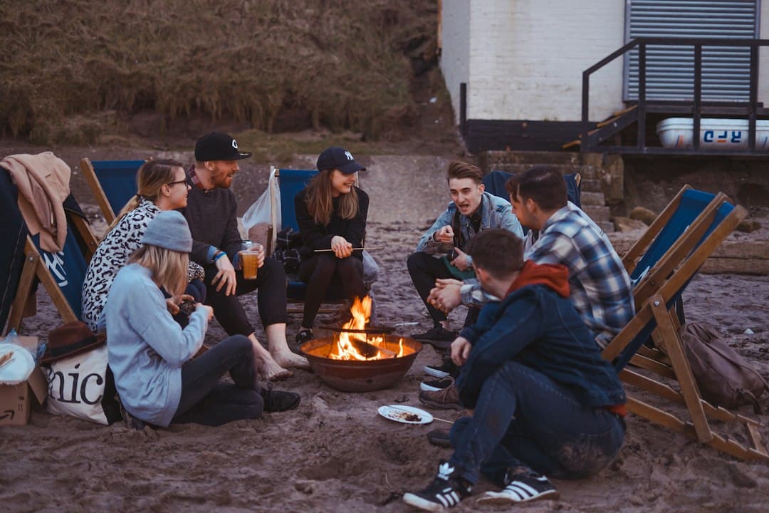 Groupe de personnes diverses socialisant autour d'un feu de camp sur la plage au coucher du soleil, assis sur des chaises longues.