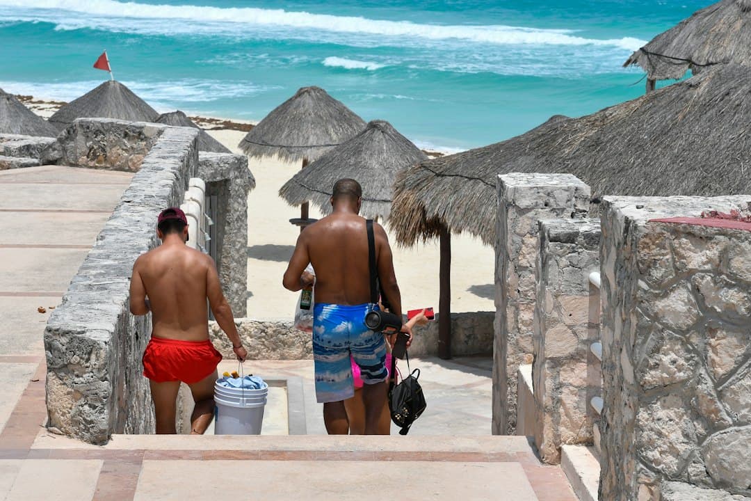 Man with a backpack walking toward a Mexican beach with palapas and turquoise water.
