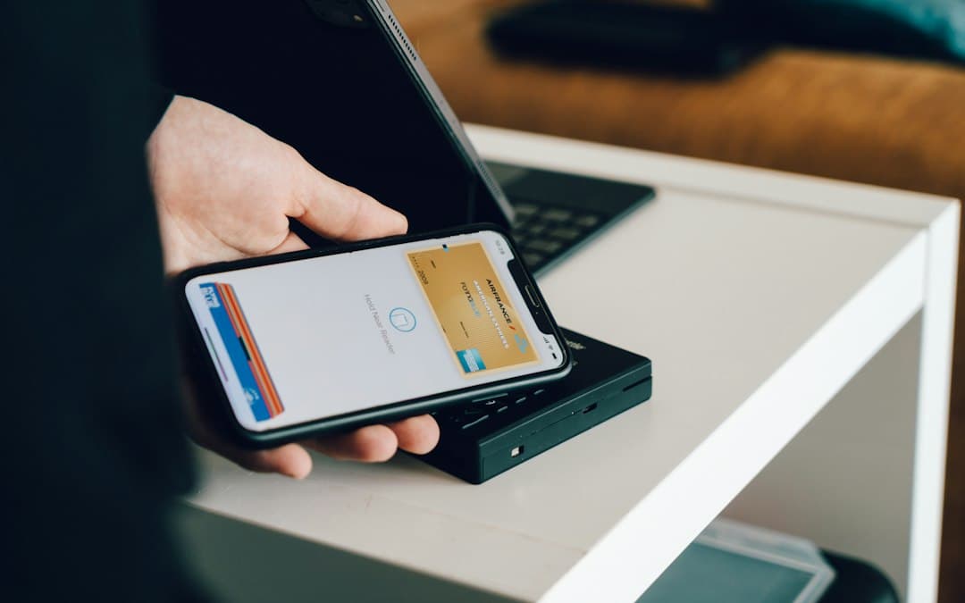 Hand holding a smartphone with a digital wallet and a gold card displayed, laptop on the desk behind.