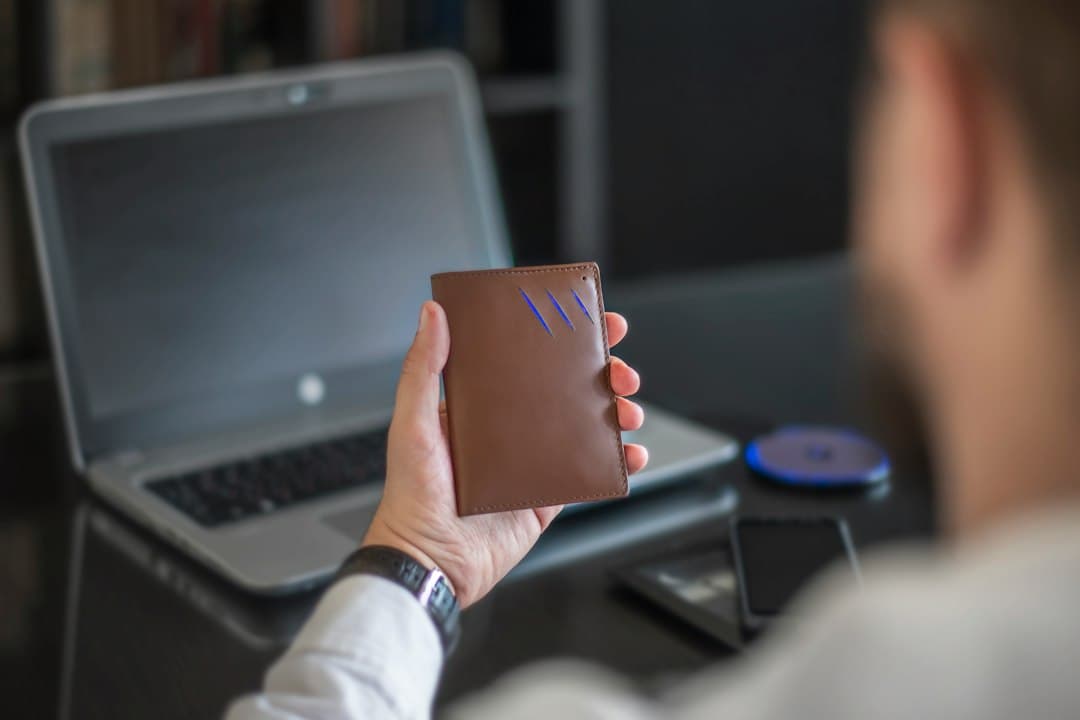 Hand holding a brown passport holder with a laptop in the background.