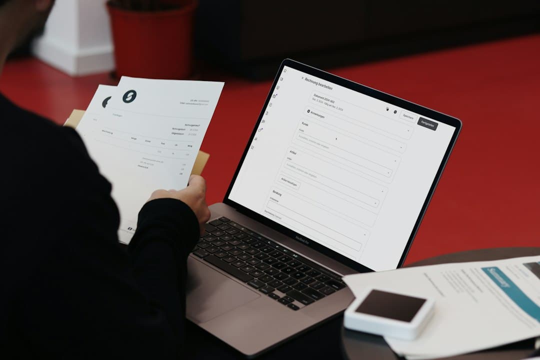 Person filling out a tax form on a laptop, with printed documents and papers on a red desk.