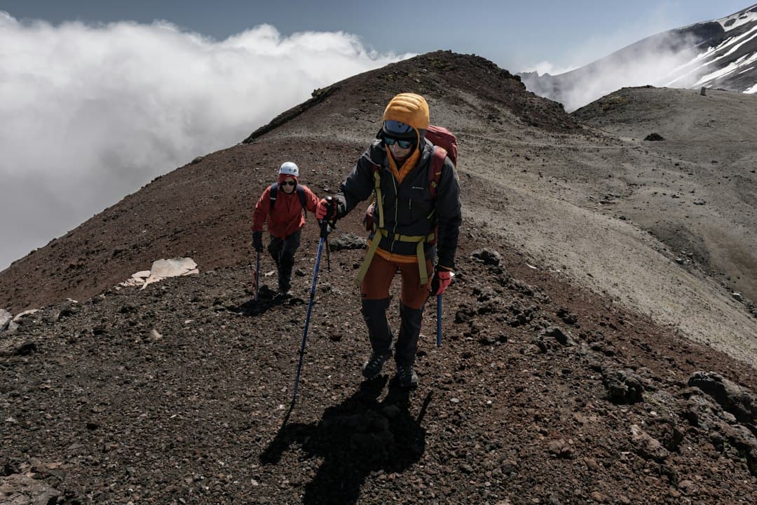 Wandelaars met wandelstokken die een vulkanische helling beklimmen, met wolken en bergtoppen op de achtergrond.