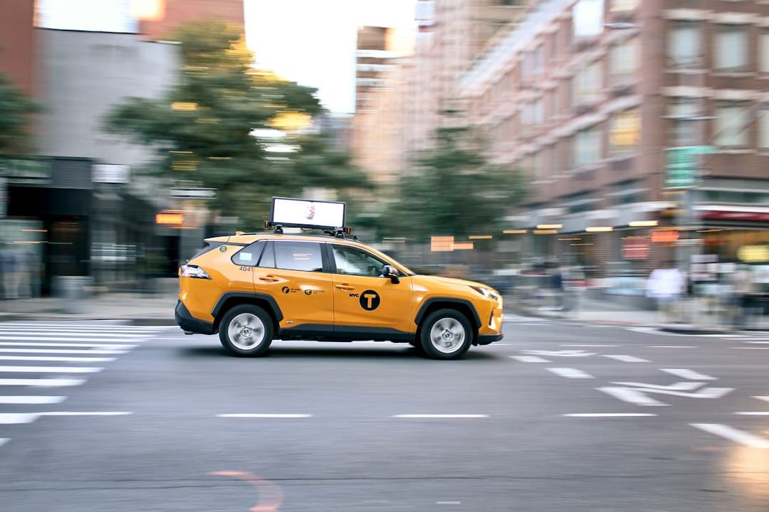 Yellow taxi driving on a busy urban street with buildings and a visible crosswalk.