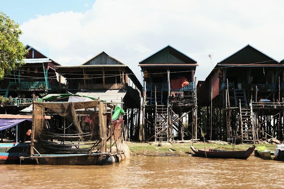 Traditional stilt houses line a brown river with fishing boats in Cambodia.